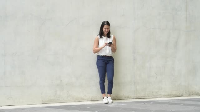 Full-Body-Shot-Of-Happy-Young-Beautiful-Woman-Leaning-On-Wall-While-Using-Phone-Outdoors