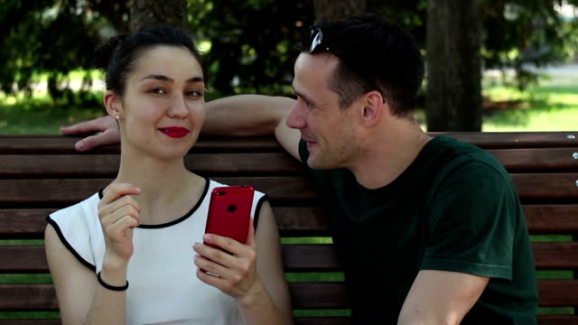 A-young-beautiful-girl-and-her-handsome-young-boyfriend-watch-the-news-in-the-social-network-on-their-smartphone-while-sitting-on-a-park-bench.