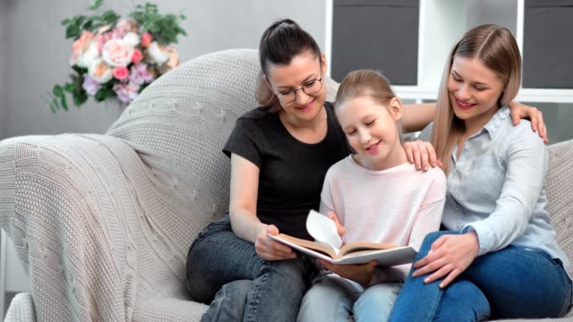 Dos-jóvenes-felices-sonriendo-del-mismo-sexo-madre-leyendo-libro-con-su-hija-en-el-interior-doméstico