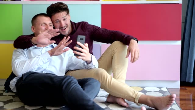 Gay-couple-using-phone-on-arm-chair-together.-Waving-bye-bye.