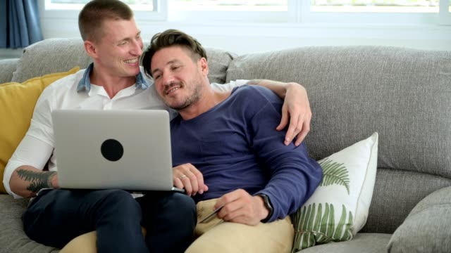 Gay-couple-relaxing-on-couch-using-laptop-computer.-Happy-shopping-and-kissing.