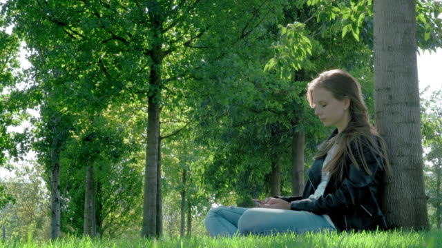 Young-beautiful-girl-in-street-clothes-in-a-park.-Holding-a-smartphone-in-his-hands,-sitting-on-the-grass-under-a-tree.-Reads-boring-message-and-photos,-Yawns-heavily.-Not-enough-sleep,-wants-to-sleep