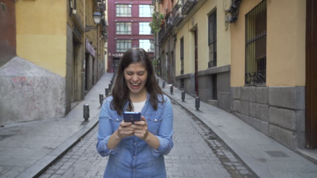 Happy-attractive-young-woman-using-smart-phone-texting-message-and-chatting-calling-friends-while-walking-on-city-street-urban-background.