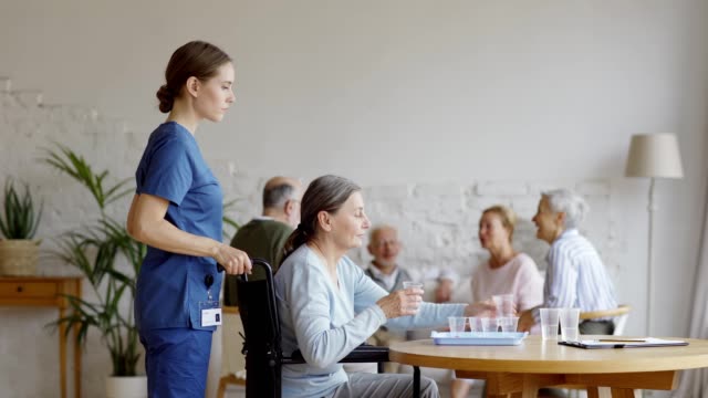 Female-nurse-pushing-wheelchair-with-disabled-elderly-woman.-Senior-patient-taking-pills-and-glass-of-water-on-table-and-joining-friends-talking-on-sofa-in-common-room-of-nursing-home