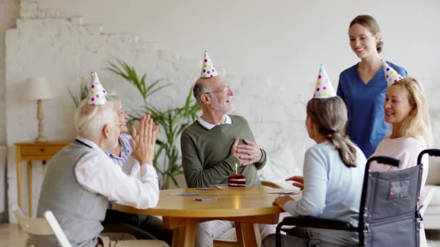 Junge-Krankenschwester-bringt-Stück-Geburtstagstorte-für-Senior-Mann-Spielen-Karten-mit-betagten-Freunden-in-Party-Hüte-in-betreutem-Wohnen.-Glücklicher-alter-Mann-empfängt-Wünsche-und-Umarmungen-und-bläst-Kerze-aus