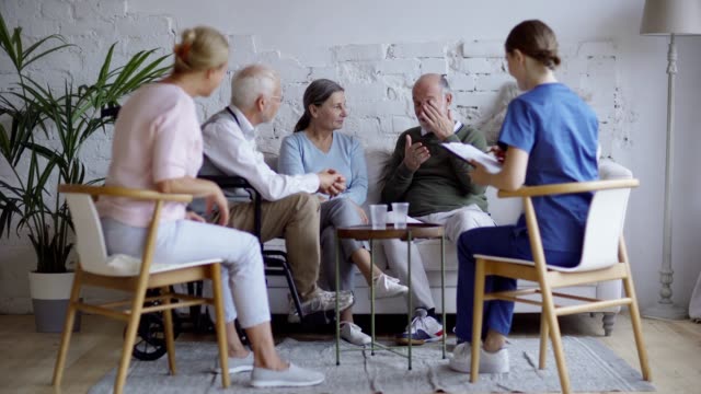 Group-of-four-senior-people-talking-in-common-room-of-nursing-home.-Young-nurse-in-uniform-or-therapist-taking-notes-in-document-on-clipboard