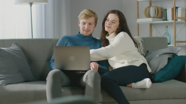 Bonita-pareja-joven-utilizar-ordenador-portátil,-divertirse,-se-ríe,-mientras-se-sienta-en-el-sofá-en-el-acogedor-apartamento.-Parejas-comprando-en-Internet,-usando-redes-sociales,-viendo-videos-y-streaming-de-contenido