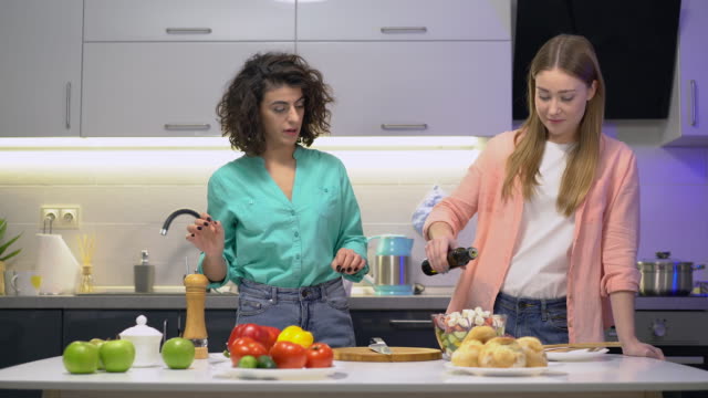 Two-attractive-women-cooking-salad-at-home,-adding-salt-and-olive-oil,-culinary