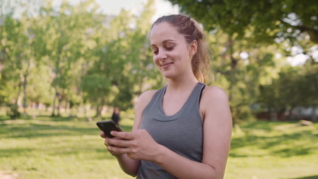 Kaukasier-fit-junge-Frau-lächelnd-beim-SMS-auf-Handy-im-Park-am-Morgen-an-einem-sonnigen-Tag