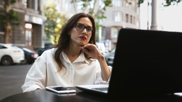 Businesswoman-in-glasses,-white-shirt.-She-sitting-at-table-with-laptop-and-smartphone-in-an-outdoor-cafe.-Cars-passing-by.-Close-up,-slow-motion