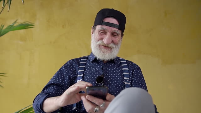 Front-view-of-handsome-high-spirited-70-aged-beard-man-in-stylish-cap-which-sitting-near-yellow-wall-and-using-his-mobile
