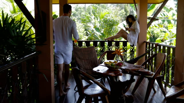 Hombre-hacer-jugo-celular-inteligente-de-uso-de-joven-sentado-en-el-balcón,-feliz-pareja-hablando-en-terraza-de-verano