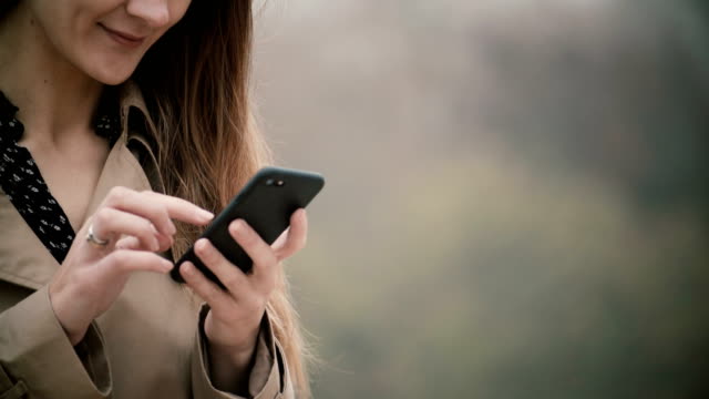 Close-up-view-of-female-hands-holding-the-mobile-phone-with-touchscreen.-Young-woman-using-smartphone-in-foggy-park