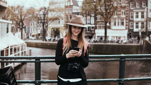 Pretty-woman-using-smartphone-on-a-bridge.-4K.-Young-lady-in-stylish-hat-with-long-hair-and-camera-types-a-message