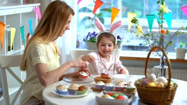 Young-mother-and-her-little-daughter-wearing-funny-rabbit-ears-cooking-Easter-cupcakes