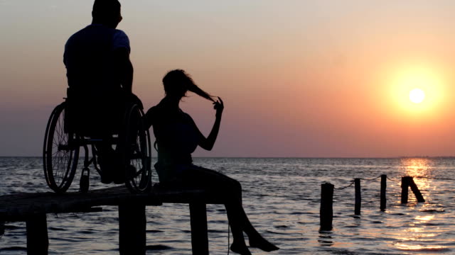 lachende-Mädchen-sitzen-auf-Pier-und-im-Gespräch-mit-Mann-im-Rollstuhl-Kulisse-von-Meer-und-Sonnenuntergang-deaktiviert