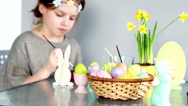 The-beautiful-girl-paints-the-figure-of-the-Easter-bunny.-There-are-paints-on-the-table,-a-basket-with-Easter-eggs-and-a-bouquet-of-daffodils