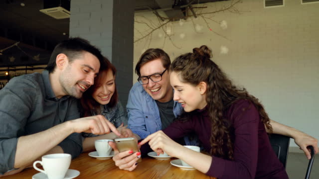 Young-people-are-watching-smartphone-screen-discussing-photos-and-laughing-during-lunch-break-in-cafe.-Friendship,-modern-technology-and-modern-lifestyle-concept.