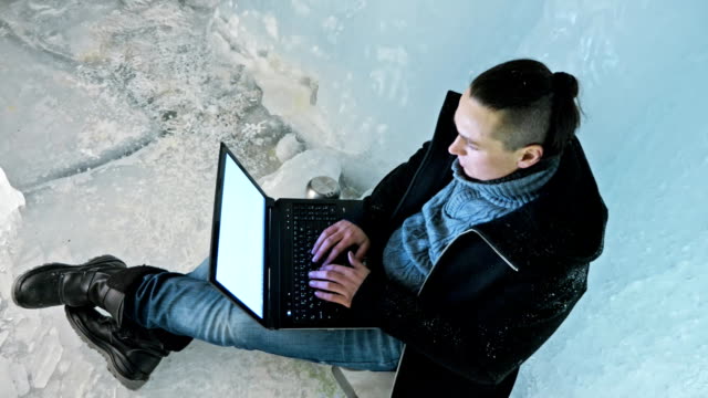 Man-is-sit-on-the-internet-in-laptop-in-an-ice-cave.-Around-the-mysterious-beautiful-ice-grotto.-User-communicates-in-social-networks.-He-crazy-strange-hipster-in-black-coat-and-fashionable-hairdo.