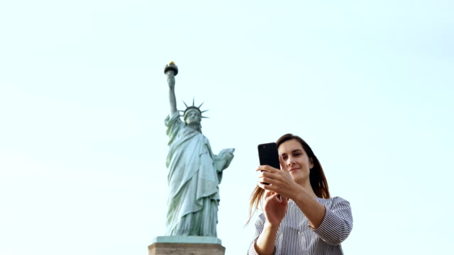 Schöne-glückliche-kaukasischen-touristischen-Frau-nimmt-ein-Selfie-Foto-mit-Smartphone-an-der-Freiheitsstatue-in-New-York-City