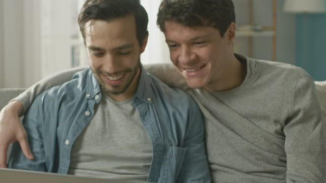 Adorable-Male-Gay-Couple-Spend-Time-at-Home.-They-Sit-on-a-Sofa-and-Use-the-Laptop.-They-Watch-Funny-Online-Videos.-Partner-Puts-His-Hand-Around-His-Lover.-Room-Has-Modern-Interior.