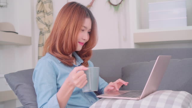 Porträt-von-schöne-attraktive-Asiatin-mit-Computer-oder-Laptop-mit-einer-warmen-Tasse-Kaffee-oder-Tee-auf-dem-Sofa-liegend-beim-Entspannen-im-Wohnzimmer-zu-Hause.-Lifestyle-Frauen-Wohnkonzept.