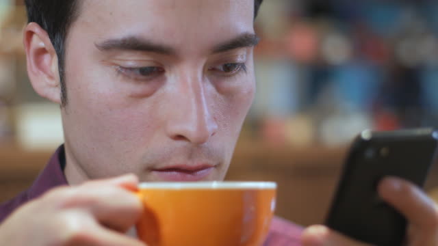 Ein-attraktive-gut-aussehender-hübscher-Hispano-Amerikaner-lateinamerikanischen-junger-Mann-ist-mit-seinem-Handy-und-trinken-eine-Tasse-Kaffee-in-einem-Coffee-Shop,-extrem,-Nahaufnahme