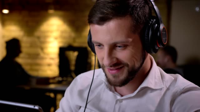 Closeup-portrait-of-young-cheerful-caucasian-businessman-listening-to-music-in-the-headphones-and-using-a-tablet