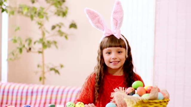 Ein-Mädchen-in-Kaninchenohren-schaut-hinter-den-Tisch,-auf-dem-ein-Korb-mit-Ostereiern-steht.