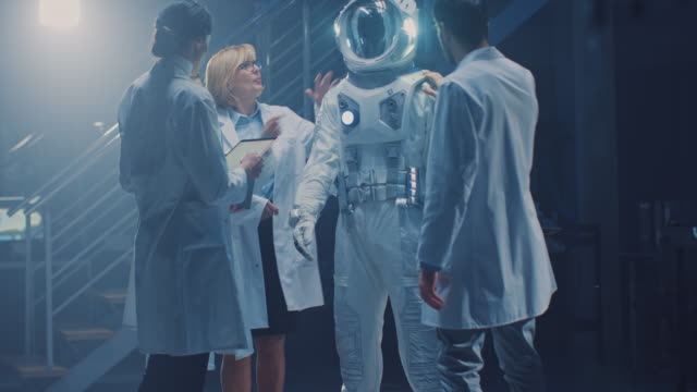 Diverse-Teams-von-Luft-und-Raumfahrt-Wissenschaftlern-und-Ingenieuren,-die-weiße-Mäntel-tragen,-haben-Diskussionen,-Benutzung-von-Computern,-Design-New-Space-Suit,-die-für-Galaxy-Exploration-und-Reisen-angepasst-sind.-Astronaut-bauen-Anzug