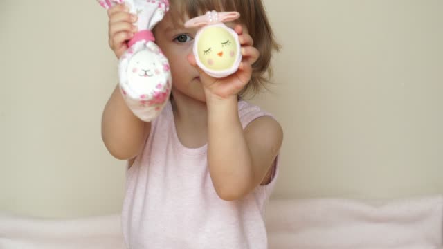 Portrait-of-cute-smiling-baby-girl-shows-chicken-eggs-in-hands,-decorated-for-Easter-bunny-and-chick,-with-painted-muzzle.