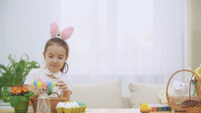 Litle-playful-girl,-wearing-bunny-ears-on-her-head-is-choosing-an-Easter-egg.-Suddenly,-somebody-noticed-her.-She-got-frightened-and-hid-down.