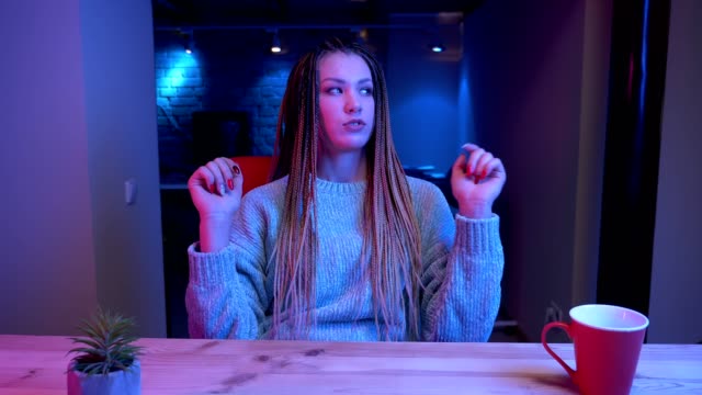 Closeup-shoot-of-young-attractive-female-blogger-with-dreadlocks-streaming-live-and-talking-cheerfully-with-the-neon-background-indoors