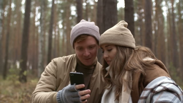 Fröhliches-Paar-mit-Smartphone-und-Chatting-im-Wald-am-Herbsttag