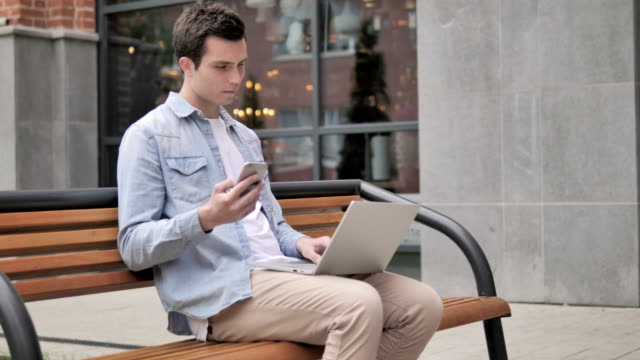 Young-Man-sitzt-mit-Smartphone-und-Laptop-im-Freien
