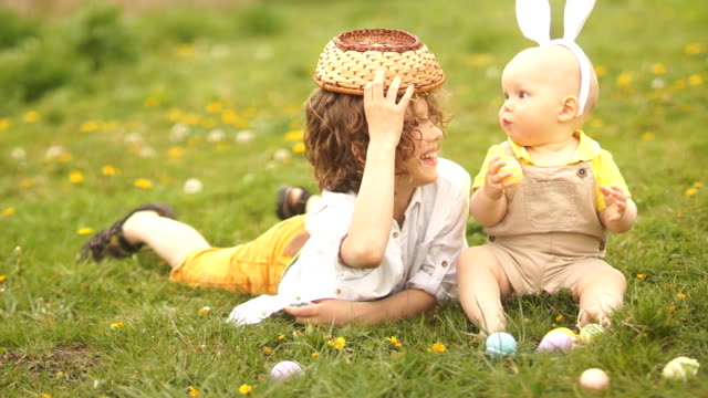Dos-hermanos,-un-adolescente-y-un-bebé-descansan-en-un-parque-en-un-claro.-Concepto-de-Pascua,-orejas-de-conejo,-cesta-de-Pascua-y-huevos.-Un-perro-corre-más-allá-de-ellos