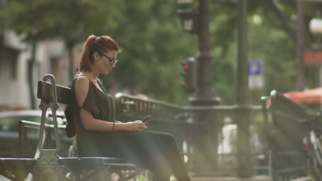 Attractive-redhead-woman-with-glasses,-freckles,-piercings-and-red-hair-writing-a-text-message-on-her-smartphone-sitting-on-street-bench,-during-sunny-summer-in-Paris.-Slow-motion.-Medium-Shot.