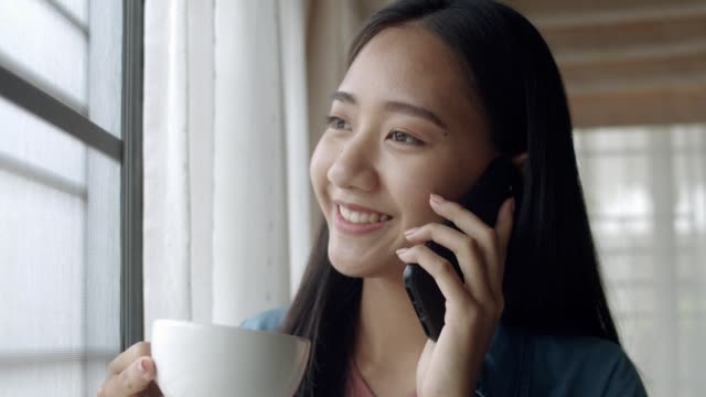 Close-up-attractive-portrait-smiling-young-asian-woman-drinking-coffee-and-talking-on-Phone-with-friends-standing-beside-window-at-home-office.
