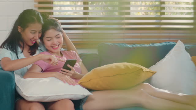 Young-Asian-Lesbian-women-couple-using-mobile-phone-check-social-media-on-internet-together-while-lying-sofa-in-living-room.