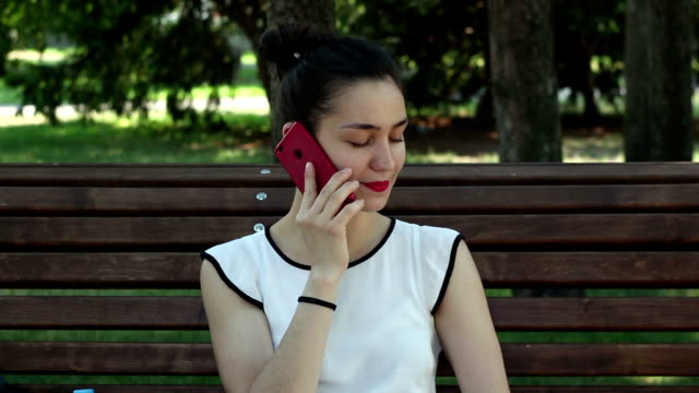 Joven-hermosa-chica-hablando-por-teléfono-mientras-está-sentada-en-un-banco-del-parque.