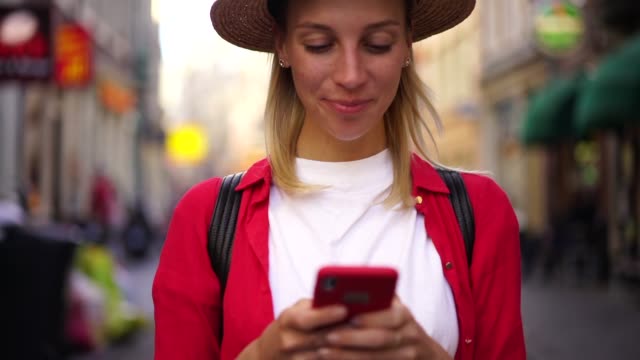 Mujer-caucásica-positiva-en-sombrero-leer-mensaje-en-el-teléfono-móvil-usando-la-aplicación-en-línea-en-la-calle-de-la-ciudad