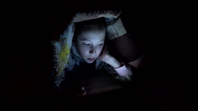 Una-niña-jugando-en-una-tableta-por-la-noche-en-una-cama-bajo-una-manta.-Vídeo-conceptual.-Primer-plano.-Vídeo-crudo.-4K.
