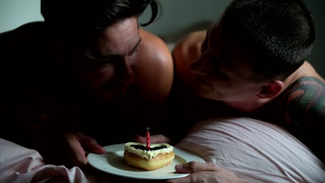 Pareja-gay-disfrutar-de-su-pastel-de-cumpleaños-juntos-en-la-cama.-Pide-un-deseo-y-sopla-la-vela-con-casi-la-oscuridad.