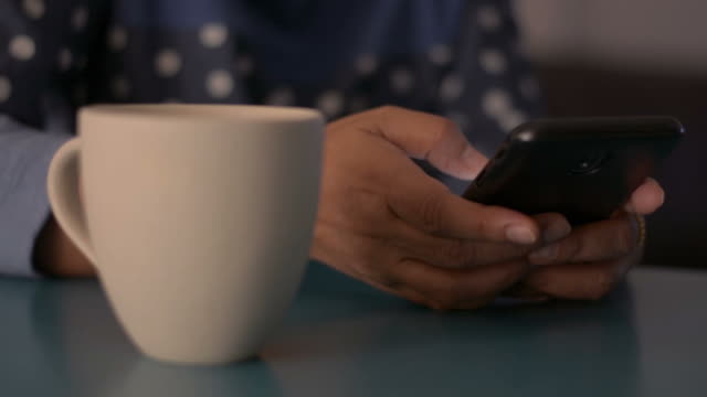 Mujer-usando-teléfono-inteligente-en-el-escritorio-y-bebiendo-una-taza-de-café-en-casa.