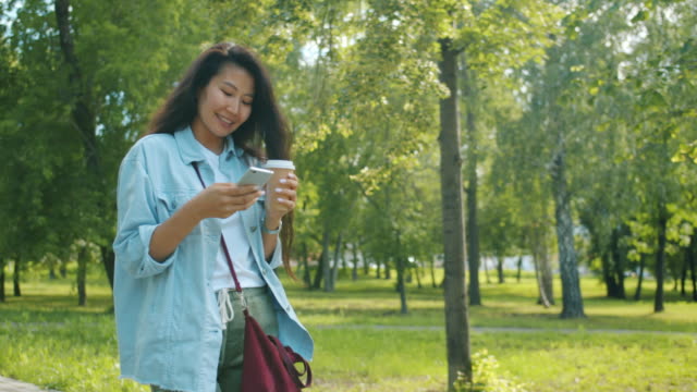 Schöne-asiatische-Frau-zu-Fuß-im-Park-mit-dem-Smartphone-und-Kaffee-lächelnd-gehen