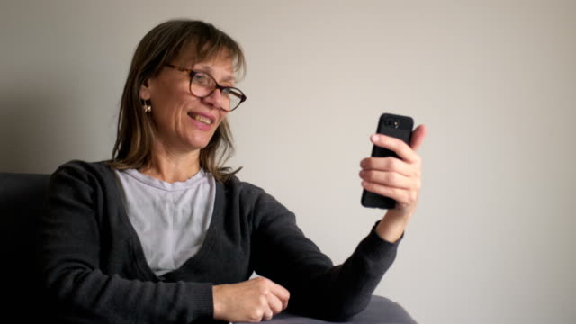 Glückliche-mittelalte-reife-Frau-hält-Smartphone-mit-Video-Chat,-mit-Social-Media-Apps-im-Telefon-sitzen-auf-der-Couch-zu-Hause,-ältere-Menschen-und-Technologie-Konzept