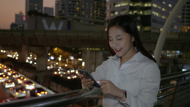 Emocionada-mujer-asiática-de-negocios-celebrando-buenas-noticias-revisando-el-teléfono-inteligente-en-el-centro-de-la-ciudad-moderna-en-la-noche-de-Bangkok-Tailandia.-Comunicación-Concept-Technology,-éxito-por-teléfono-móvil