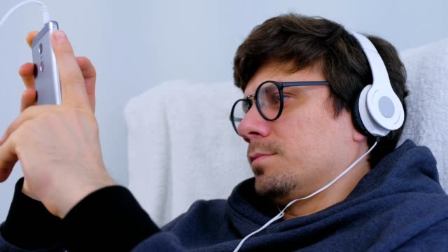 Man-in-glasses-and-headphones-watches-video-on-mobile-phone.-Side-view.