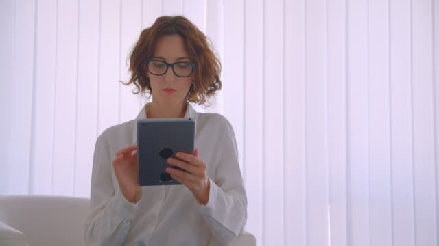 Closeup-portrait-of-adult-redhead-caucasian-businesswoman-in-glasses-using-the-tablet-looking-at-camera-sitting-in-the-armchair-in-the-white-office