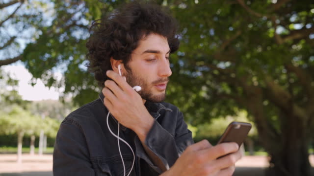 Porträt-eines-modernen-lächelnden-jungen-Mannes,-der-Kopfhörer-in-seine-Ohren-einfügt-und-online-auf-dem-Handy-im-Park-SMS-abgibt